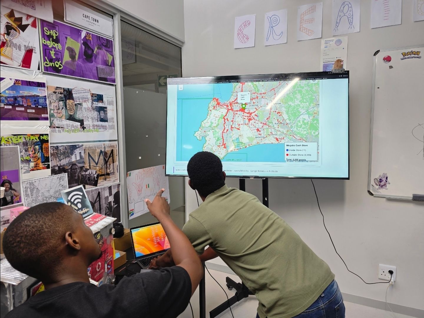 Two timbuk2 team members in the Cape Town office pointing at a wall screen showing a map of Cape Town covered in thousands of red survey points