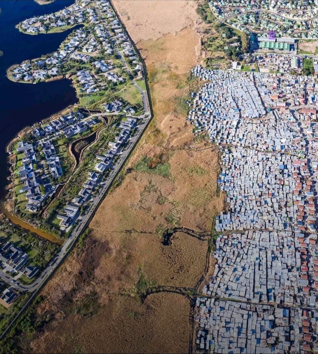 Aerial photograph of a Cape Town suburb: a lakeside estate of large houses on the left, a dense settlement of small roofs on the right, a strip of open veld between them